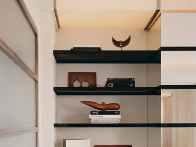Modern minimalist built-in shelving unit with dark walnut floating shelves, white cabinetry, and carefully arranged books, wooden sculptures, and ceramic objects against a neutral wall.