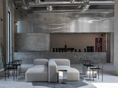 Modern industrial cafe lounge with raw concrete walls, a large rippled stainless steel feature wall, light grey modular sofas, black metal chairs, and exposed ceiling ducts.