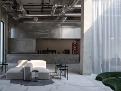 Spacious industrial-style cafe lounge with polished concrete floors, exposed ductwork, a large reflective stainless steel feature wall, white modular boucle sofas, black metal chairs, and a curved green velvet sofa in the foreground.