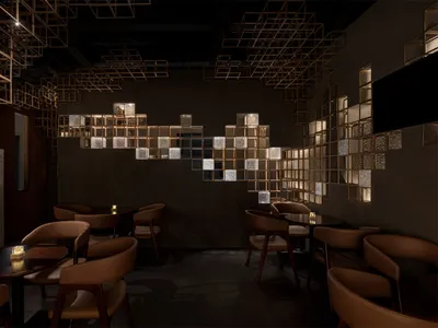 Dimly lit modern restaurant interior featuring brown leather dining chairs, dark round tables, and a striking illuminated brass cube grid art installation covering the back wall.