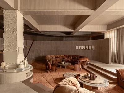 Modern living room featuring a large curved terracotta velvet sofa, a prominent white textured plaster column, layered circular coffee tables, and warm earthy tones throughout the space.