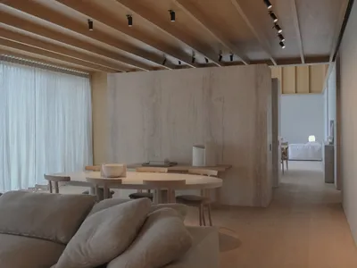 Modern minimalist dining room featuring a large curved light-wood table, matching wooden chairs, a prominent raw concrete partition wall, exposed wood beam ceiling with track lighting, and a soft beige sofa in the foreground.