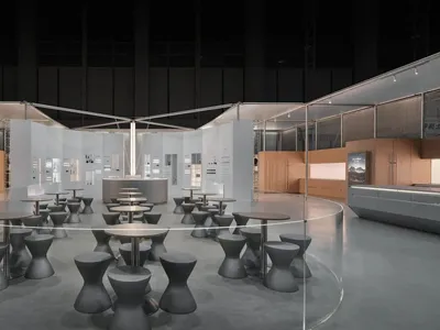 Modern exhibition space with rows of dark concrete hourglass-shaped stools and round tables inside a large circular glass partition, set in an industrial-style hall with concrete floors and wooden display walls.