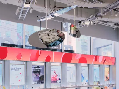 Modern cosmetics store interior featuring a large curved grey display island, red accent wall with circular patterns, product testing stations, and large windows with city views.