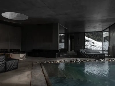 Modern brutalist indoor swimming pool with black leather modular sofas, marble tables, and large glass windows revealing a snowy mountain forest outside.