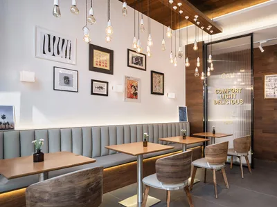 Modern cafe interior with grey upholstered banquette seating, light walnut dining tables and chairs, multiple hanging Edison bulb pendant lights, and a gallery wall of framed art.
