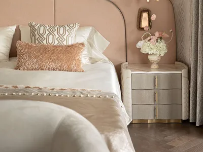 Modern luxury bedroom with blush pink curved upholstered headboard, textured beige wallpaper, white satin bedding, rose gold fur pillow, and elegant gold pendant light beside a marble nightstand with fresh flowers.