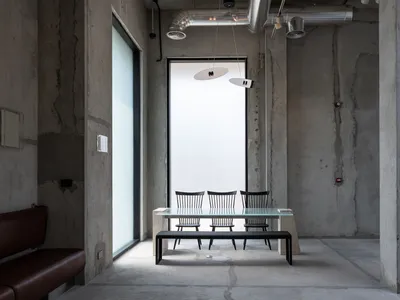 Industrial-style dining area featuring raw concrete walls, a glass dining table with black spindle-back chairs, a black bench, and two modern pendant lights suspended from an exposed ceiling with visible ductwork.