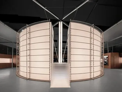 Modern art installation with two large curved fabric screens in off-white with metal frames, a central staircase leading upward, set in a spacious minimalist gallery with dark ceiling and concrete floor.