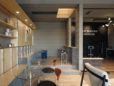 Modern cafe and livehouse interior featuring a long service counter with light birch plywood upper cabinets, blue glass mosaic tile wainscoting, round glass tables with molded plywood chairs, and a raised performance area in the background.