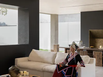 Woman wearing colorful traditional Taiwanese Indigenous dress and headdress sitting on a white sofa in a modern minimalist concrete living room with large windows.