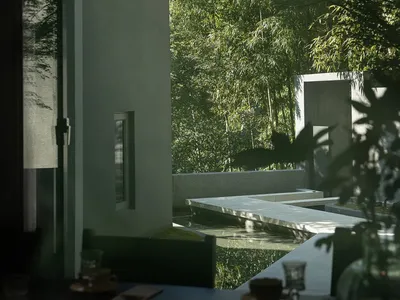 Modern minimalist dining area with dark table set, looking out through large openings to a concrete courtyard with water pond and dense bamboo forest.