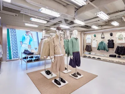 現代極簡服裝精品店室內空間，裸露混凝土天花板、工業日光燈照明、鋼鐵展示架，以及穿著當代休閒服的模特兒站在米色地毯上。