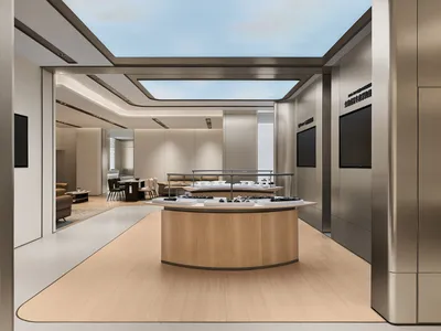 Modern luxury jewelry boutique interior featuring a large curved oak display island, brushed stainless steel walls, light wood flooring, and a large rectangular skylight revealing blue sky.