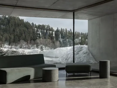 Modern minimalist living room with concrete walls and floor, featuring a curved green fabric sofa, matching ottoman, cylindrical concrete side tables, and a massive picture window overlooking a snowy forest and mountain.