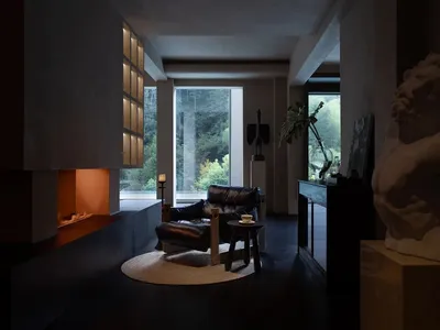 現代簡約客廳，設有內嵌式混凝土壁爐與溫暖爐火，黑皮革休閒椅放置於圓形黃麻地毯上，前景有大型大理石人體雕塑，落地窗外可見茂密綠林。