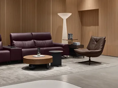 Modern living room with a burgundy leather sofa, brown leather swivel chair, round walnut coffee table, and vertical wood slat wall with a tall white floor lamp.