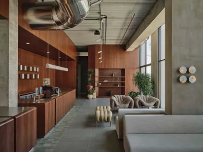Modern industrial cafe lounge with extensive walnut wood cabinetry, concrete walls, large windows, boucle armchairs, and a prominent stainless steel ventilation duct suspended from the ceiling.