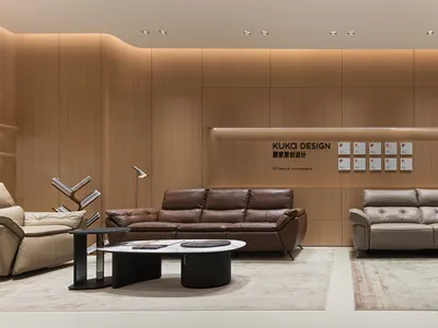 Modern living room display with a brown leather sofa and a light beige leather sofa arranged on separate area rugs in front of a curved wood-paneled wall with recessed lighting and awards.
