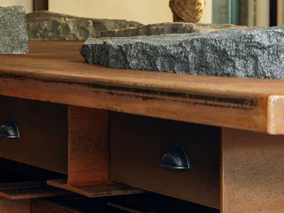 Close-up of a custom bathroom vanity featuring a thick weathered steel frame with integrated drawers, topped by a massive rough-hewn stone basin, resting on a natural stone base.
