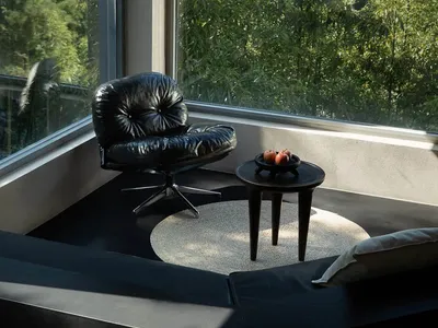 Modern black leather lounge chair with chrome base placed on a circular jute rug next to a dark wood side table holding red apples, set in a sunlit corner with floor-to-ceiling windows overlooking dense green bamboo forest.