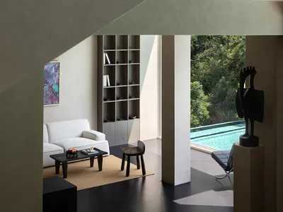 Modern minimalist living room with raw concrete architecture, white sofa, dark wood furniture, built-in shelving, abstract art, and a large opening revealing a swimming pool surrounded by lush greenery.