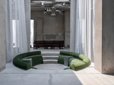 Modern brutalist interior featuring a large curved emerald green velvet sofa arranged in a sunken circular platform, surrounded by sheer white curtains and exposed concrete walls with visible ductwork.