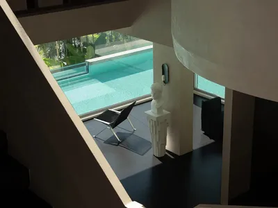 View from above of a modern curved concrete staircase overlooking a contemporary living area with a white classical bust on a pedestal, black leather chair, and large glass windows revealing an infinity pool surrounded by tropical greenery.