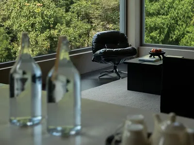 Modern living room interior with a black leather lounge chair placed beside floor-to-ceiling windows overlooking a dense green forest, with a low black side table and foreground table setting.