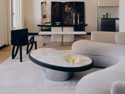 Modern minimalist living and dining area featuring a curved white marble coffee table, cream bouclé sectional sofa, black sculptural chair, and a large four-panel black lacquered folding screen with gold artwork.
