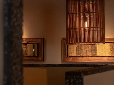 客廳溫馨內景，大型竹編圓筒吊燈、粗獷石材牆面、木框織品藝術品，以及大地色系的棕色與米色抱枕。