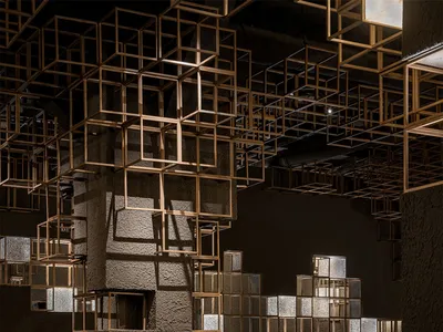 Interior of a stylish restaurant featuring textured concrete columns, brown leather dining chairs, round tables, and an elaborate ceiling installation of illuminated golden cubic metal frames.