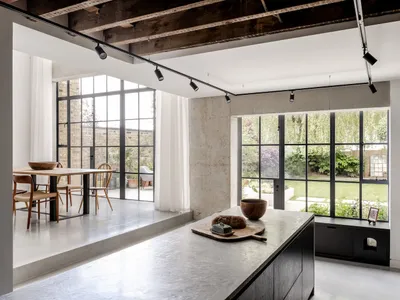 Modern industrial-style kitchen with polished concrete island, dark wood ceiling beams, black steel-framed windows overlooking a garden, and wooden dining table in the background.