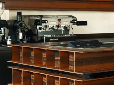 A sculptural coffee table made from stacked rusted H-beam steel sections sits on a dark tiled floor in front of a professional espresso machine.