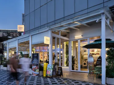Modern cafe named CALI with large glass windows, white structural frames, outdoor seating, and potted plants along a patterned stone pedestrian street at dusk.