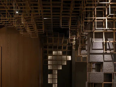Modern restaurant bar interior featuring an intricate brass cube grid ceiling installation, textured concrete walls, and a long dark bar counter with backlit circular stone inlays and leather stools.
