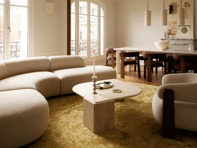 Spacious Parisian living and dining room featuring a large curved white boucle sofa, irregular marble coffee table, mustard-yellow rug, and three large white mushroom-shaped pendant lights hanging from a high ceiling with ornate molding.