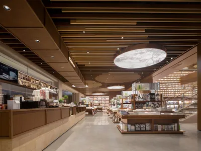 現代複合式書店咖啡廳內部，暖色木格柵天花板、巨型圓形天窗燈具、嵌入式書架，以及左側的服務吧台。