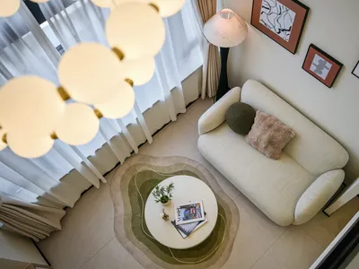 Top-down view of a modern living room with a curved cream boucle sofa, two decorative pillows, a round white coffee table on a layered green and beige organic-shaped rug, a cloud-like multi-sphere pendant light, and framed abstract art on the wall.