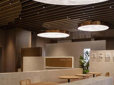 Modern café interior featuring light-colored terrazzo counters, wooden tables, rattan chairs, and large circular pendant lights with leaf-patterned projections on the curved wooden slat ceiling.