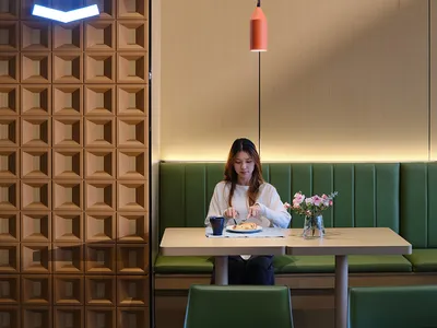 Woman dining alone at a light wood table in a modern restaurant booth with green upholstery, a coral pendant lamp, and a striking textured wooden wall featuring a glowing neon logo.