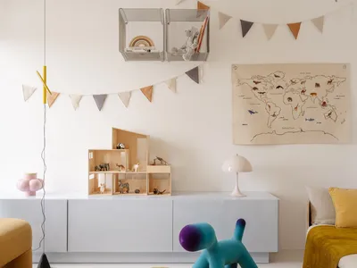 Bright and airy children's playroom with white storage, wooden dollhouse, animal figurines, cloud-shaped beige felt rugs, a blue-purple gradient dog stool, and a world map on the wall.