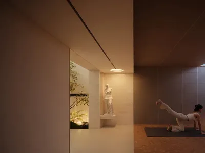 穿著白色運動服的女子在黑色瑜伽墊上練習瑜伽，背景為微水泥牆面的極簡工作室、壁龕中的古典維納斯雕像與溫暖環境照明。