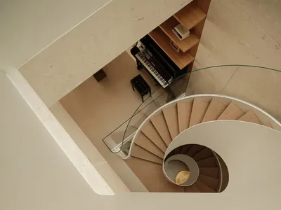 Top-down view of a modern spiral staircase made of light oak with white curved balustrade, glass railing, and a black upright piano nestled in a built-in wooden shelving nook.