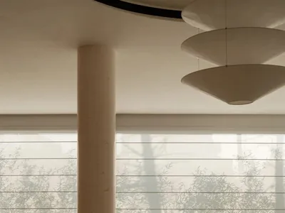 Modern dining room featuring a large round travertine table, curved upholstered chairs, a cylindrical concrete column, and a sculptural three-tiered pendant light with large windows overlooking bamboo in the background.