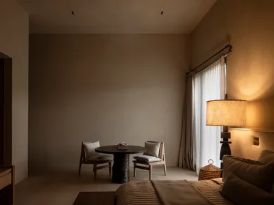 現代簡約臥室內景，深色橡木圓桌搭配兩張軟墊椅，高挑編織立燈投射溫暖光線，中性米色牆面，以及鋪有紋理亞麻寢具的床鋪。