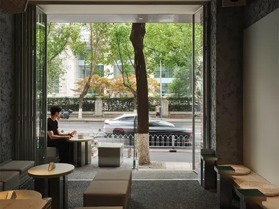 Modern minimalist cafe interior with concrete walls, wooden benches, beige ottomans, round tables, and a large open facade framing a street tree and passing cars.