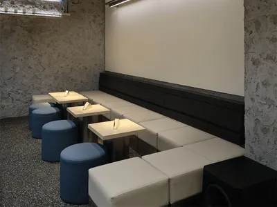 Modern restaurant interior featuring raw concrete walls, built-in white upholstered banquette, small square tables, blue cylindrical ottomans, and dramatic linear LED lighting.