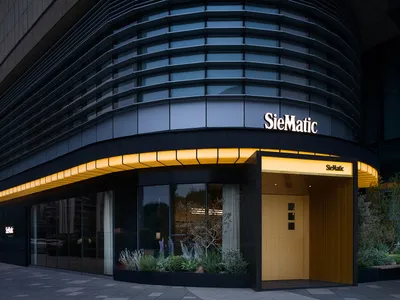 Night view of a modern luxury kitchen showroom with a curved dark glass facade, warm yellow LED lighting along the canopy, and landscaped planters at the entrance.