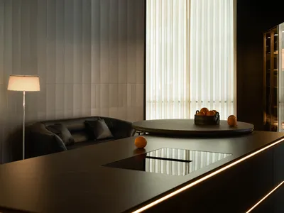Modern dark interior featuring a curved wooden ceiling, vertical grey wall panels, black leather sofa, round dining table, and a long kitchen island with integrated LED lighting and induction cooktop.